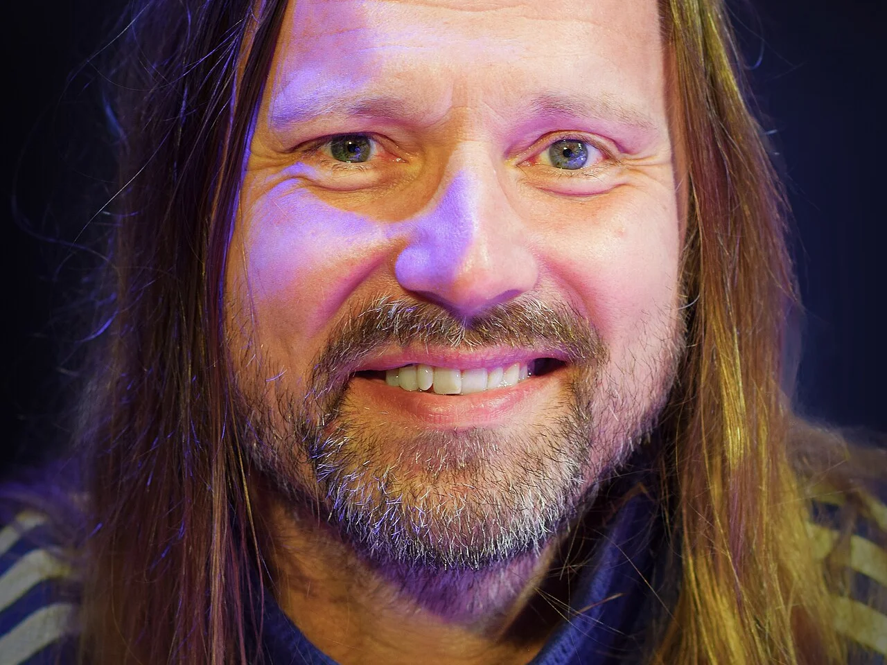 Närbild av en leende Max Martin med långt hår och skägg i lila scenljus